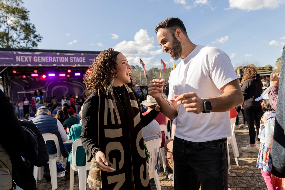 Two people smile and interact while standing near seated attendees at an outdoor event, with a stage labeled "Next Generation Stage" in the background.