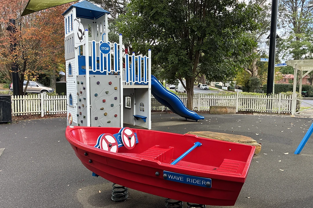 Children's playground with a red "Wave Rider" boat on springs and a climbing structure with a blue slide. Surrounding trees and a white fence add charm.