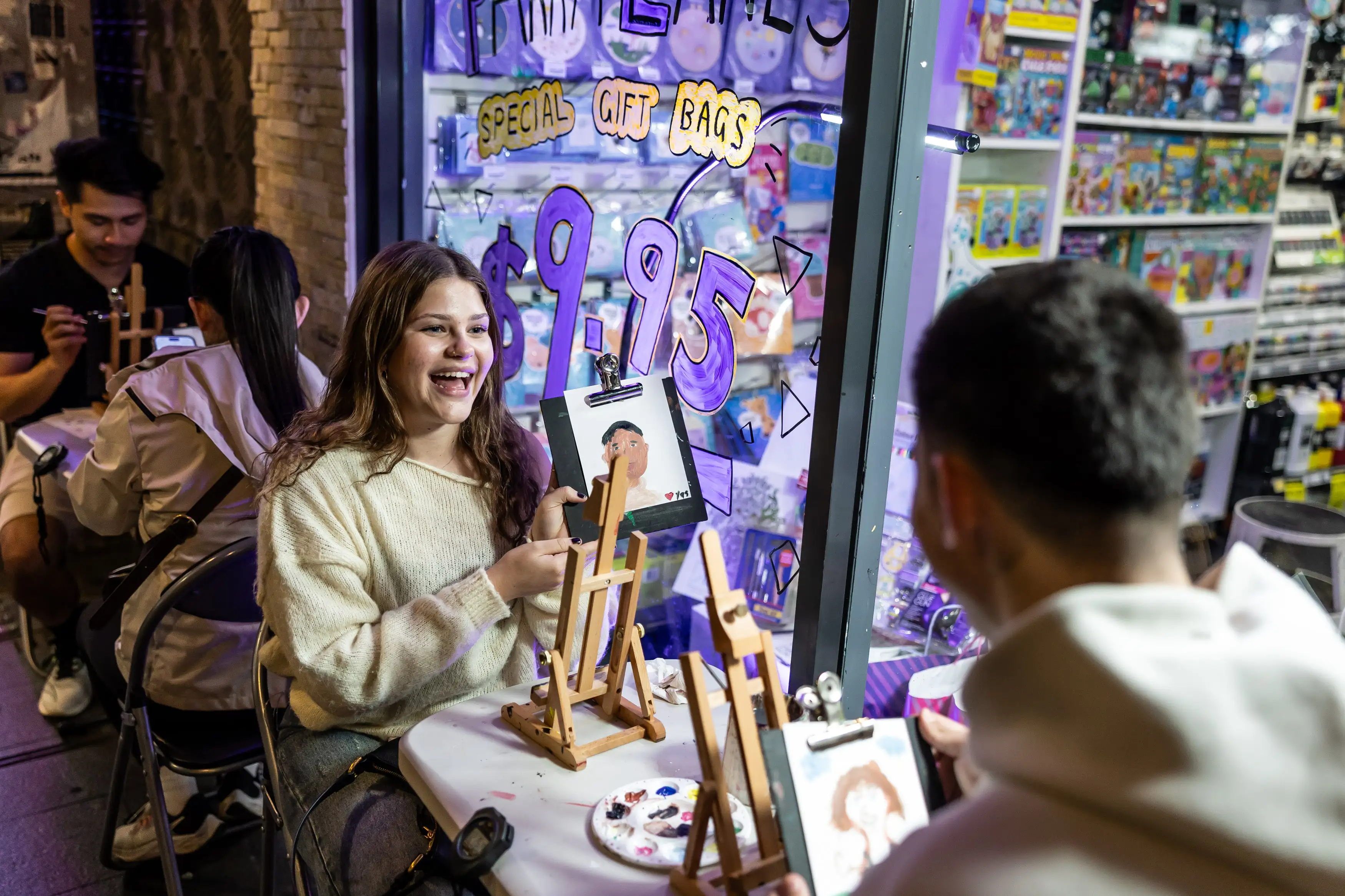 A joyful young woman paints at an art class, smiling as she sits with others at small easels inside a colorful store with a $9.95 gift bag sign.