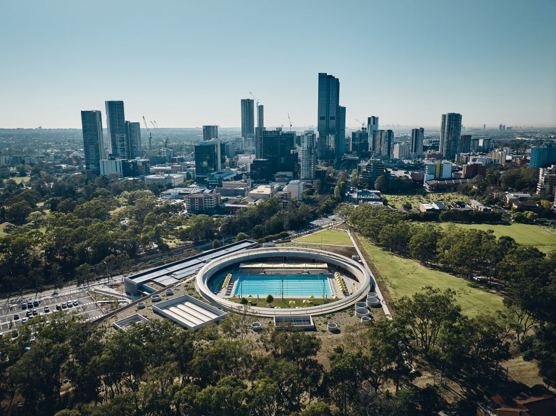Aerial view of a modern cityscape with a circular building housing a swimming pool in the foreground. Tall skyscrapers rise in the background amid greenery.