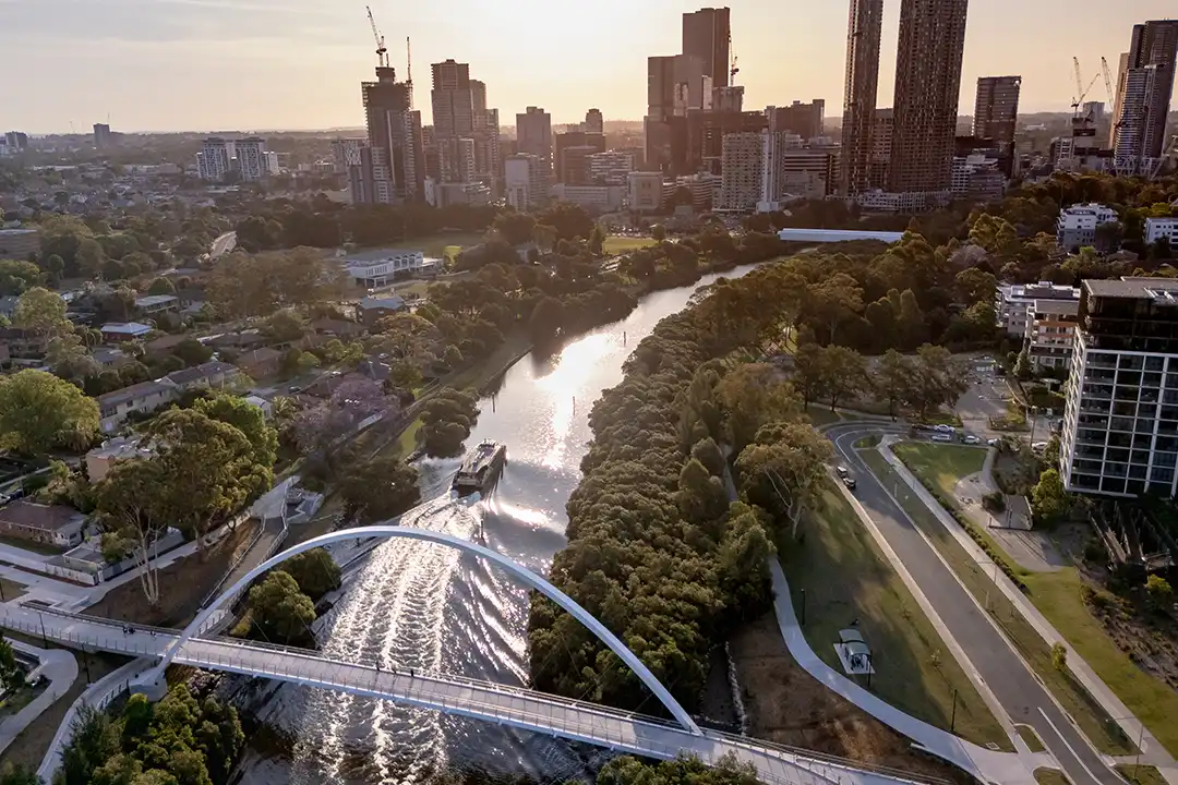 Aerial view of a river with curved footbridge, boat, and lush greenery. Modern city skyline and sunset light create a serene urban landscape.