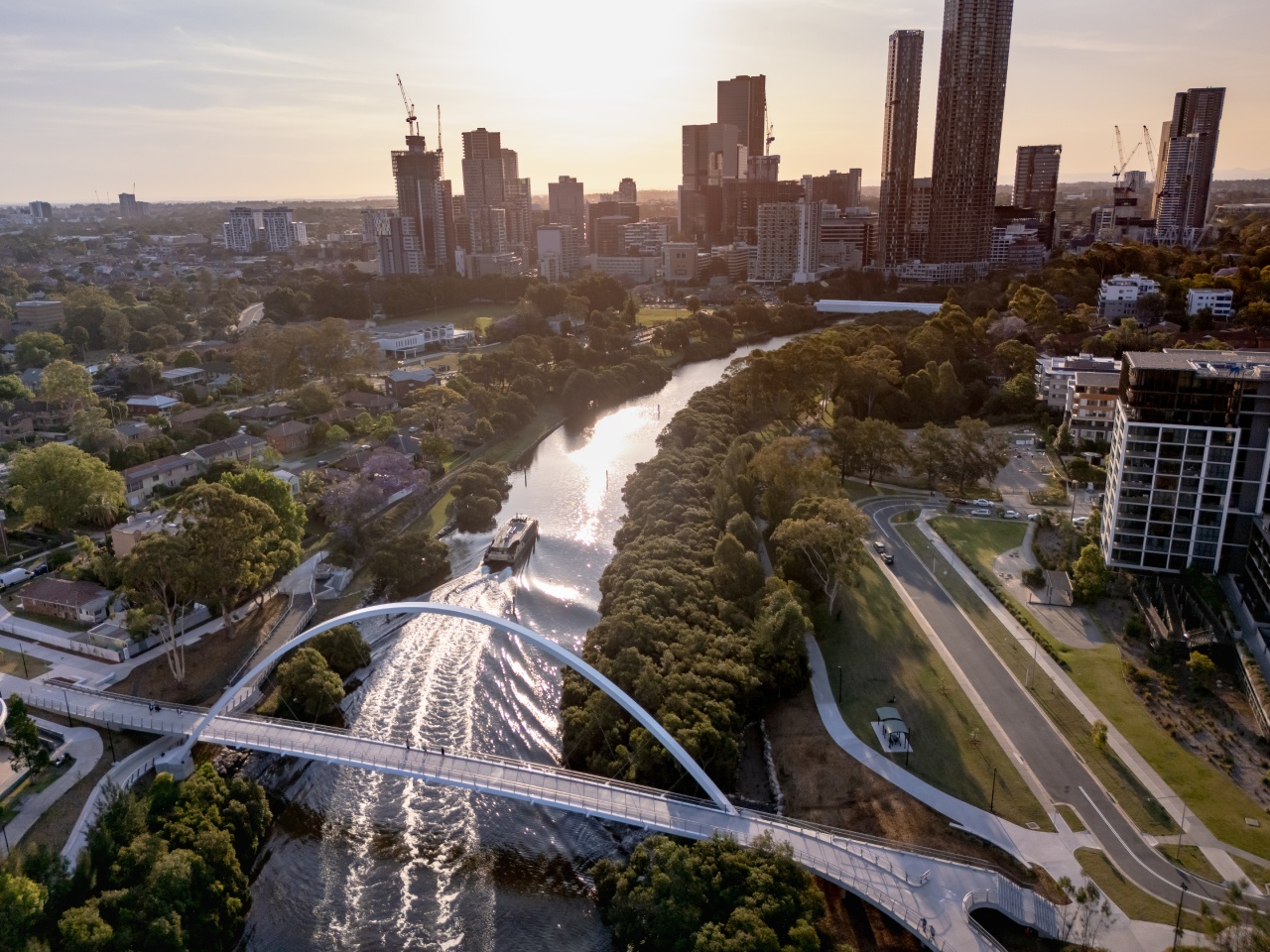 Aerial view of a cityscape at sunset, featuring a modern arched bridge over a tree-lined river and a boat.