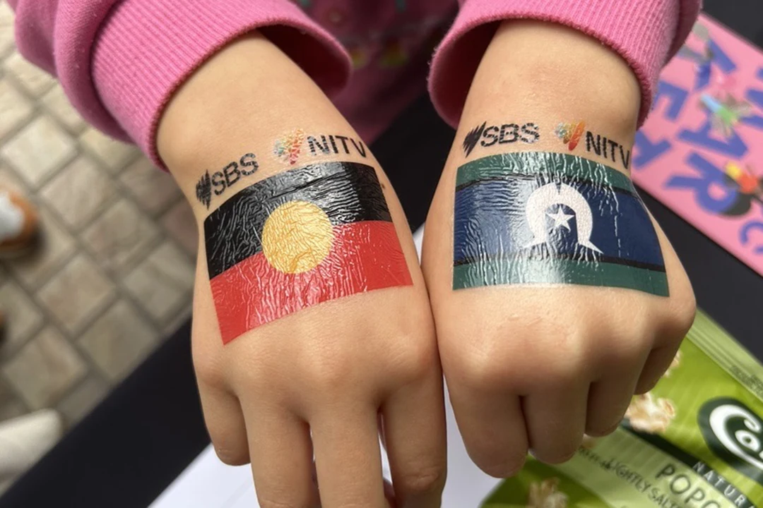 Two hands display temporary tattoos: the Aboriginal flag on the left and the Torres Strait Islander flag on the right, both labeled "SBS" and "NITV". The tone is youthful and vibrant.