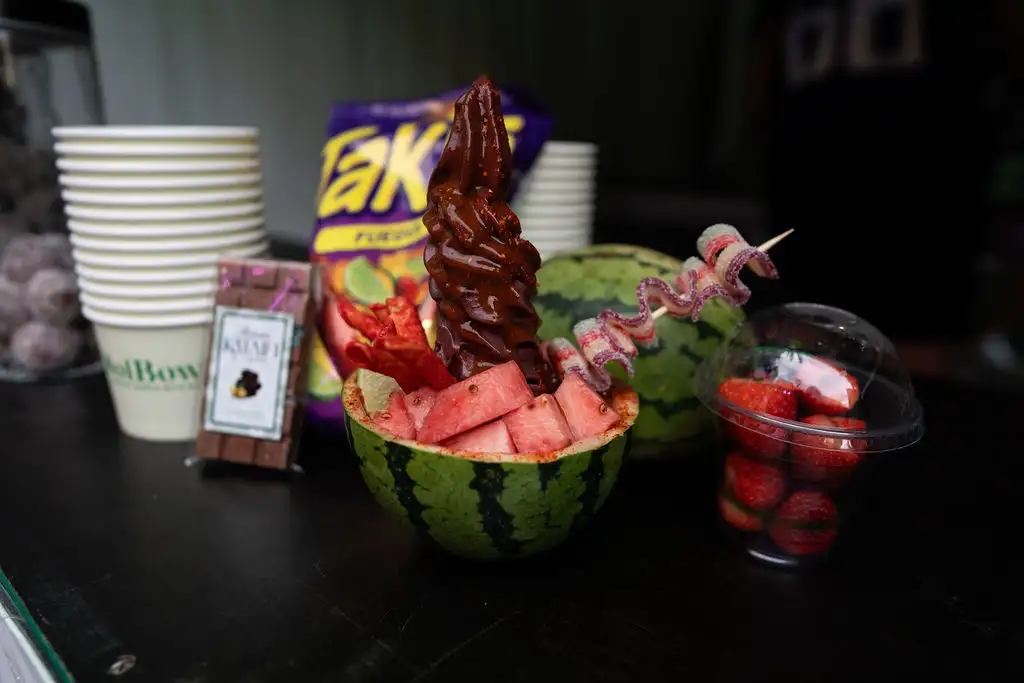 A watermelon half filled with fruit, topped with a spiral skewer, sits beside strawberry cups, Takis chips, chocolate bars, and paper cups on a table.