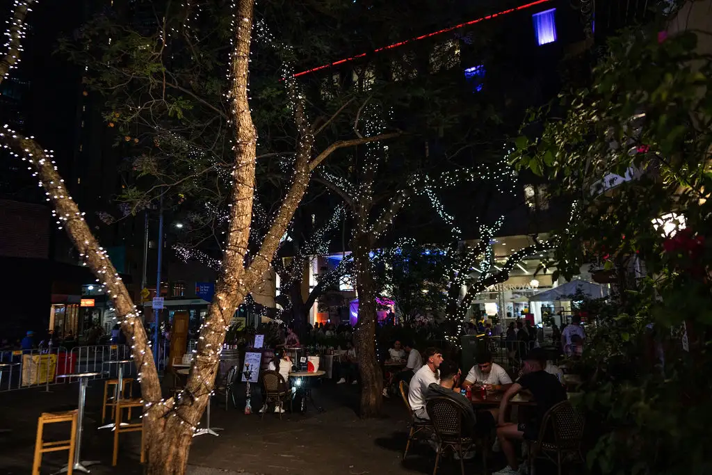 Outdoor night scene with trees adorned in fairy lights. People sit at tables, enjoying a lively atmosphere. Warm, festive, and inviting ambiance.