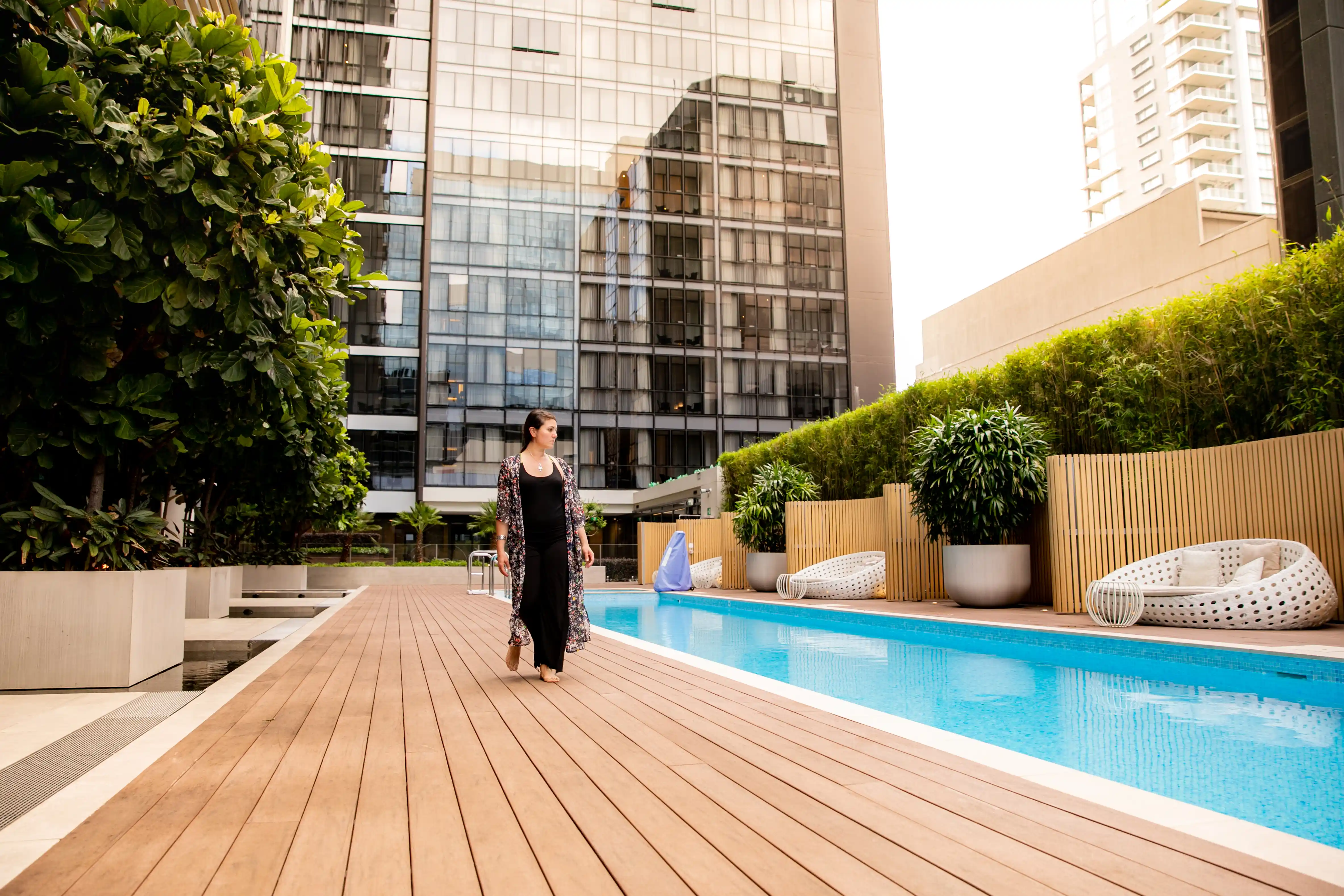 A woman in a long black dress walks by a modern rooftop pool bordered by plants and wooden fencing, with tall glass buildings in the background.