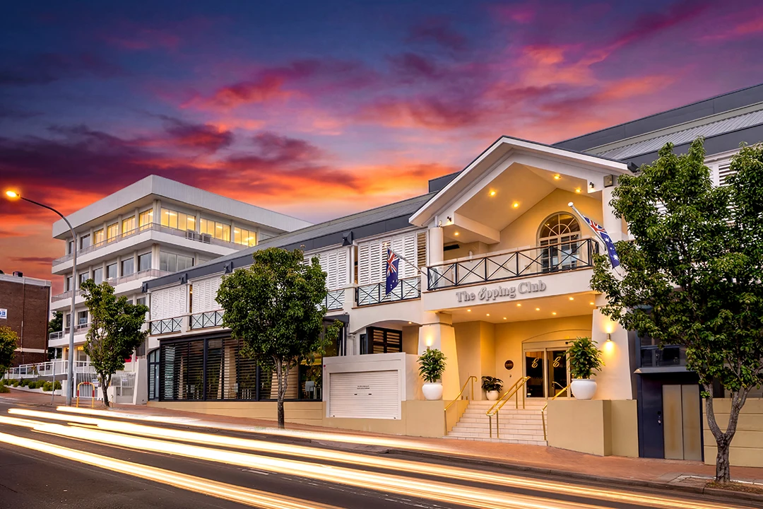 Elegant building with arched entrance, lit warmly under a vibrant sunset sky. Streetlights create streaks, adding dynamic energy to the urban setting.