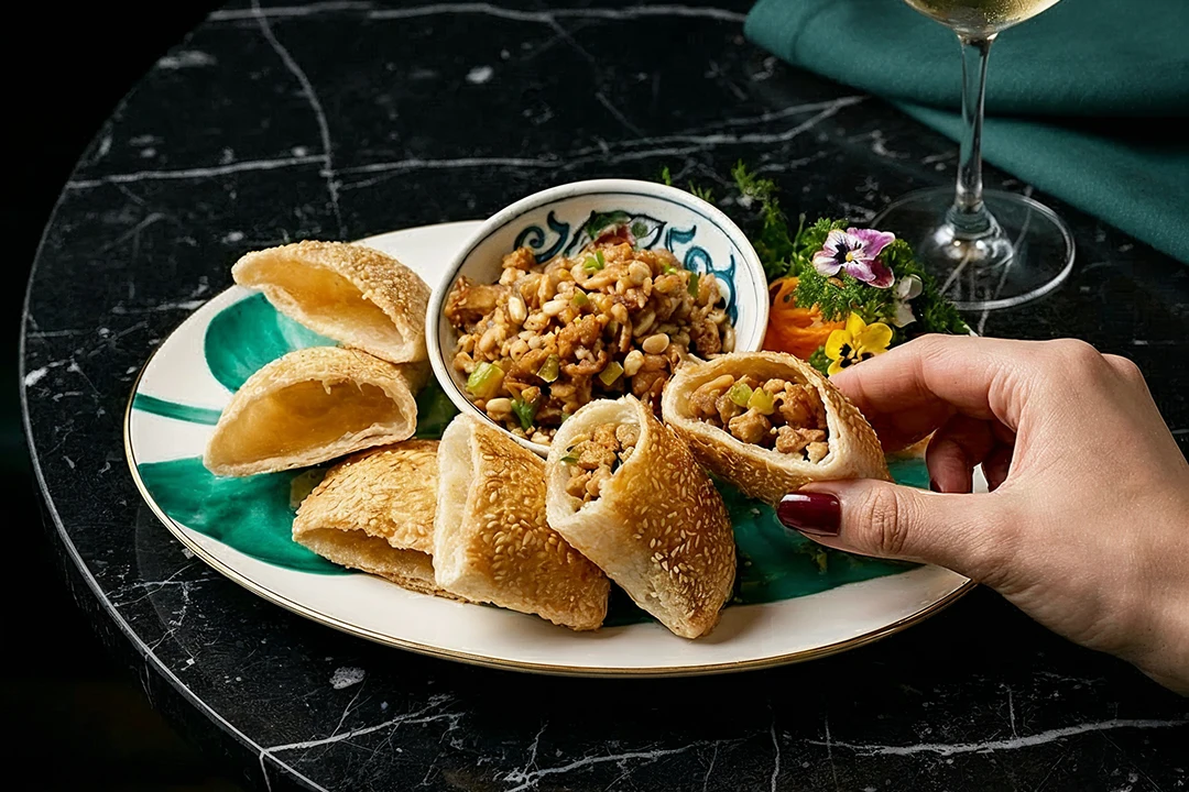 A hand with red nail polish picks up a sesame pastry filled with seasoned meat. The dish is artfully arranged on a marble table, with a floral garnish.