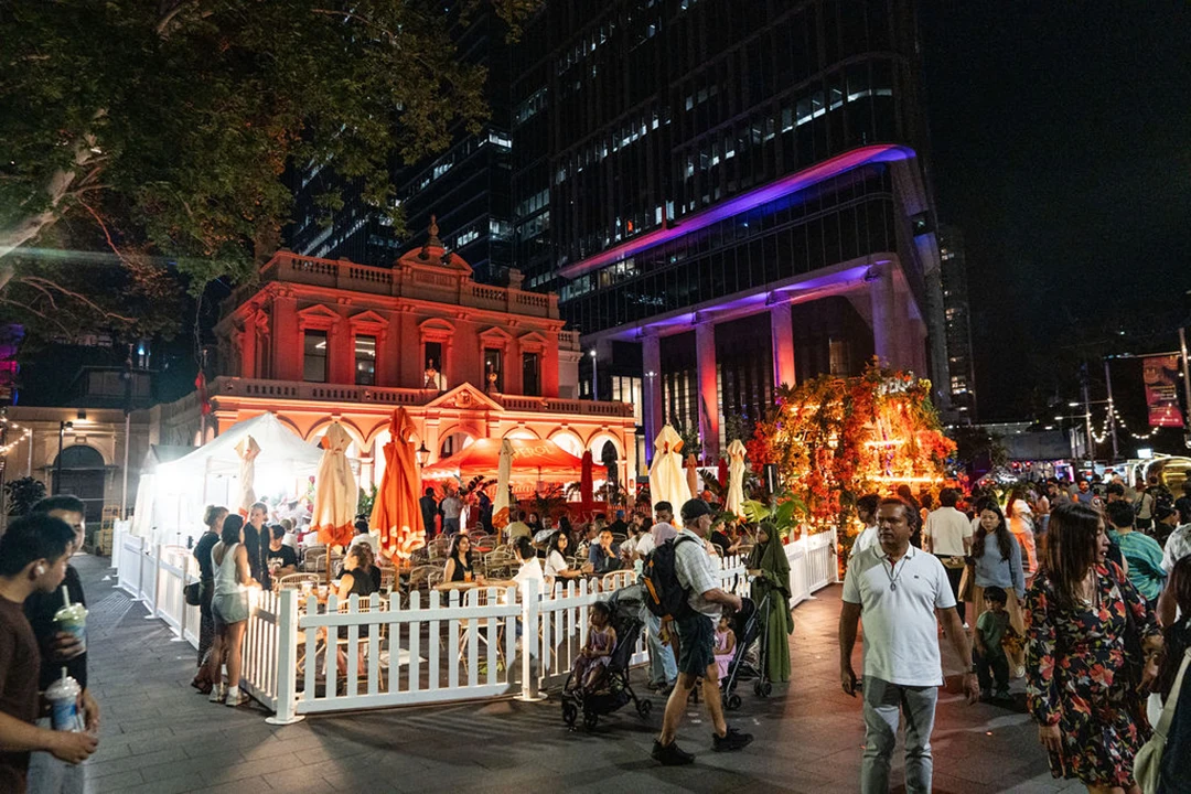 A bustling nighttime urban scene with people gathering outside a vibrantly lit building in red and orange. Festive atmosphere with colorful lights and decorations.