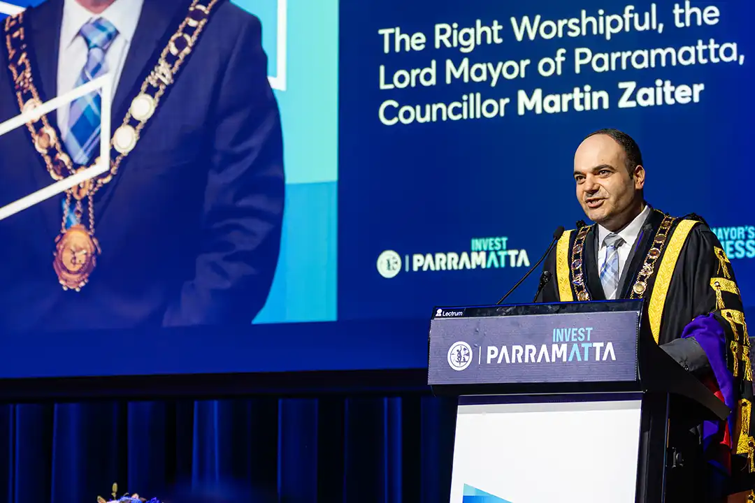 A man in ceremonial attire speaks at a podium labeled "Invest Parramatta." A large screen behind him displays his image and title as Lord Mayor.