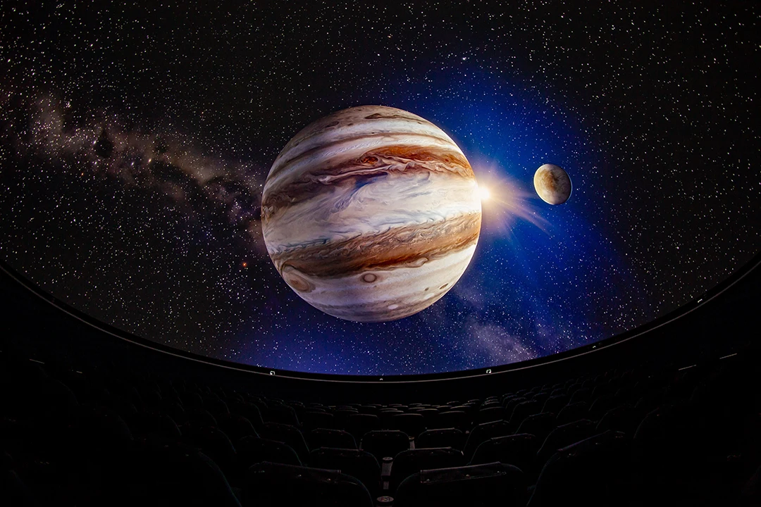 A planetarium display shows a vivid image of Jupiter and its moon against a starry backdrop. Empty theater seats in the foreground add to the immersive feel.