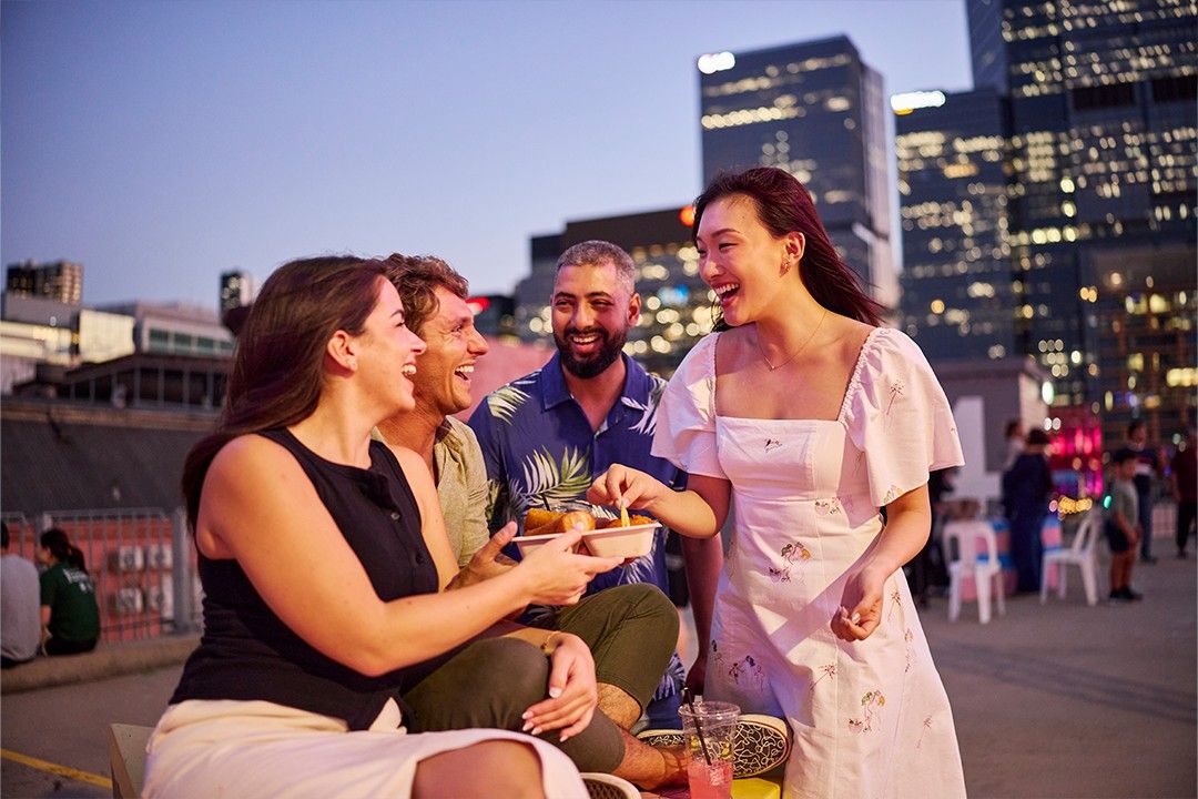 Four friends share laughter and snacks on a rooftop with a city skyline backdrop at dusk. The mood is joyous and relaxed, with warm lighting.