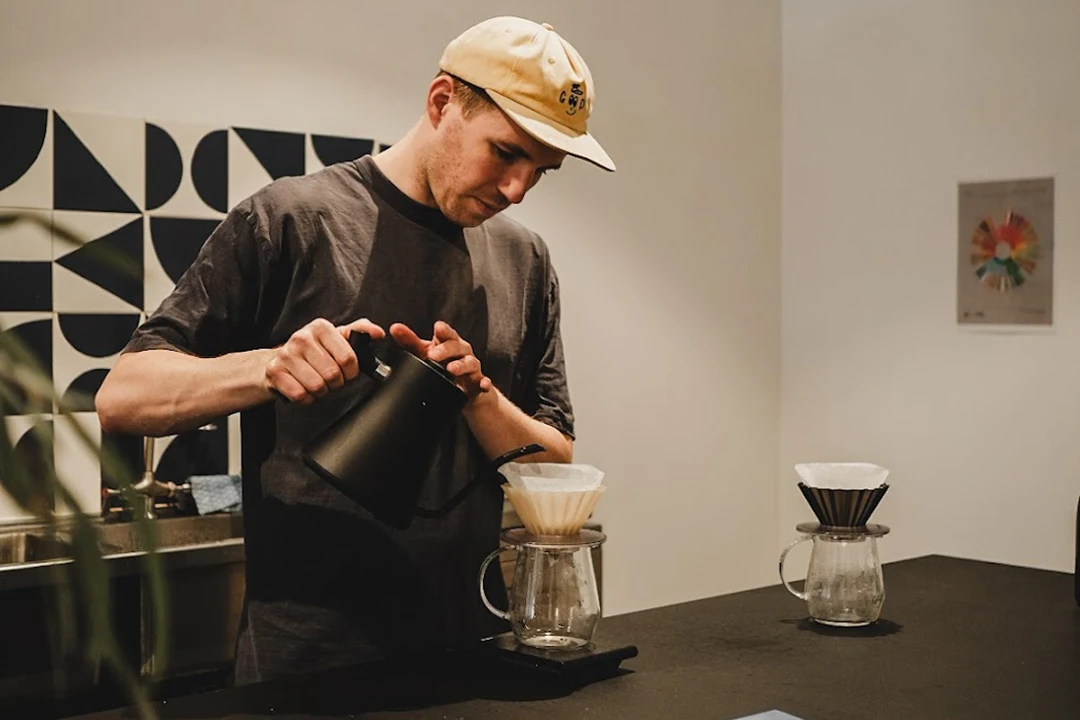 A person in a casual cap and T-shirt pours water from a kettle into a drip coffee maker on a black counter, conveying focus and calm.