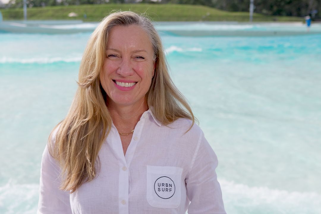 Smiling woman with long blonde hair wearing a white shirt with "URBNSURF" logo, standing in front of a bright blue wave pool, exuding a cheerful vibe.