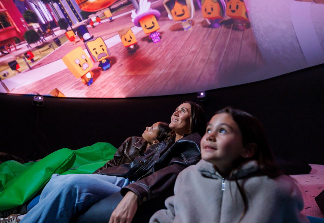 A family of three watches a colorful animated film projected on a dome screen, sitting on bean bags. They look engaged and delighted.