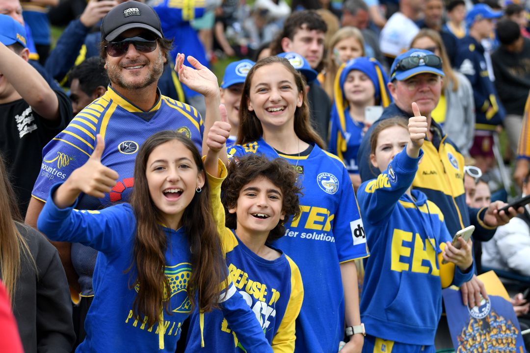Fans wearing blue and yellow sports jerseys smile and give thumbs up at a sporting event. The crowd behind them looks cheerful and energetic.
