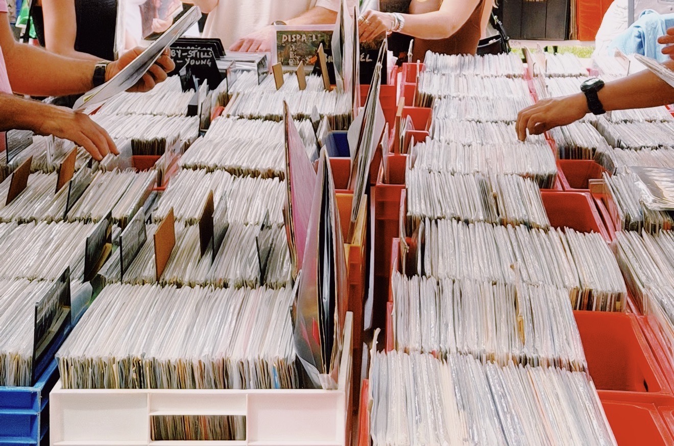 Hands browse through rows of vinyl records in colorful bins at a busy market. The scene conveys a nostalgic and lively atmosphere.