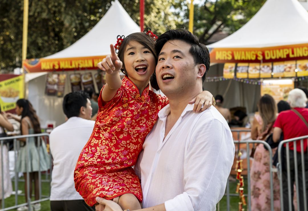 At a lively festival, a smiling father holds a young girl in a red Chinese dress as she points excitedly near Malaysian dumpling stalls.
