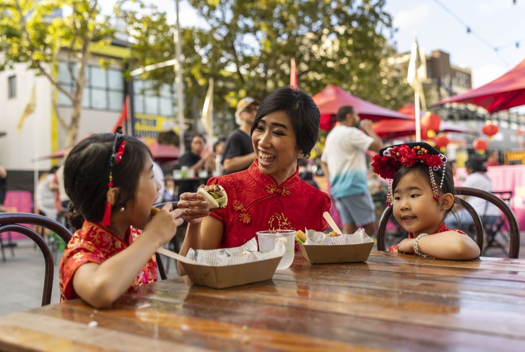 Smiling mother in a red dress shares food with two young daughters at an outdoor Asian market, creating a warm, joyful family moment.