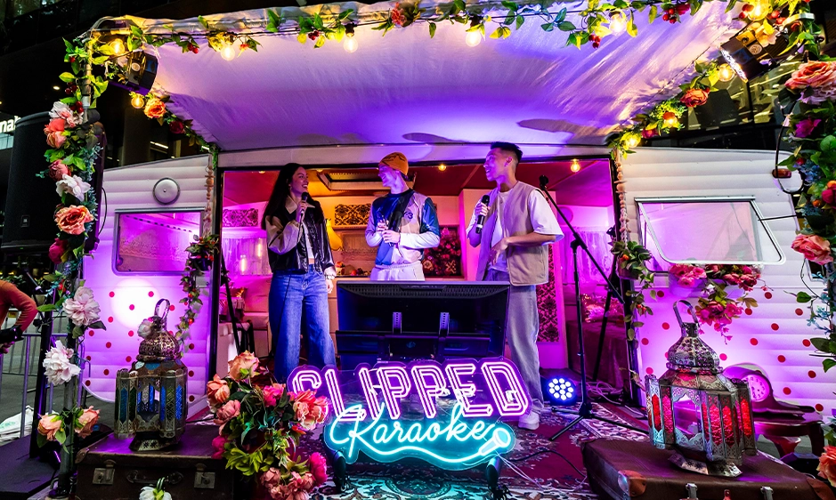 Three singers perform on a vibrant karaoke stage, framed by flowers and lanterns, under purple lighting and a glowing “CLIPPED Karaoke” sign.