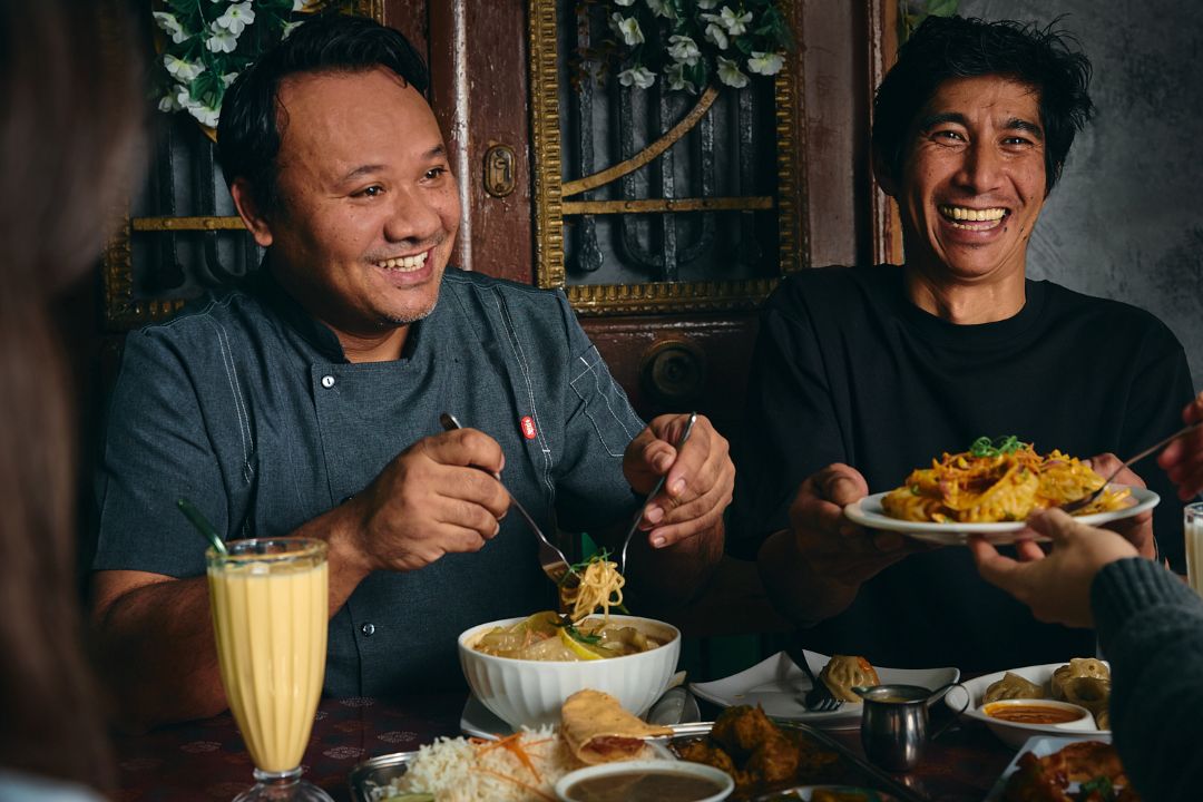 Two smiling men share a meal at a cozy restaurant table; one lifts noodles while the other presents fried rice, creating a warm, convivial mood.
