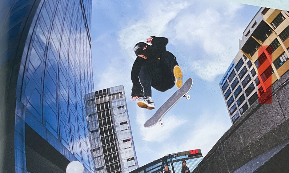 Skateboarder performs a midair trick above a city plaza, framed by glass skyscrapers and blue sky, creating an energetic urban scene.