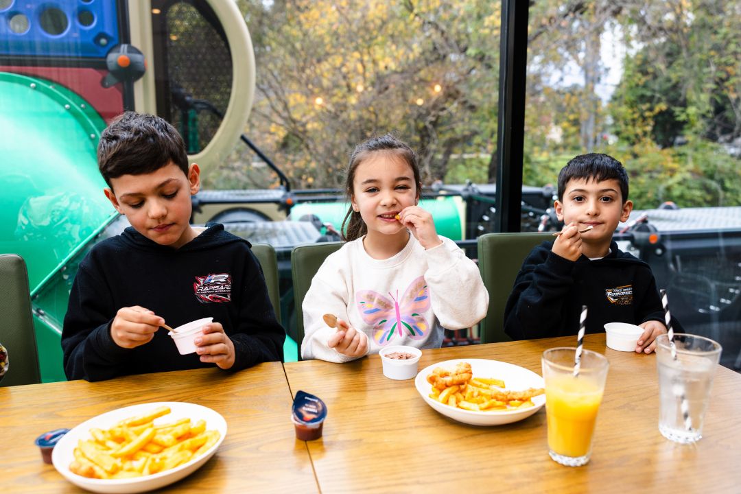 Three children enjoy fries and drinks at a restaurant table, smiling and eating, with a colorful indoor playground visible behind them.
