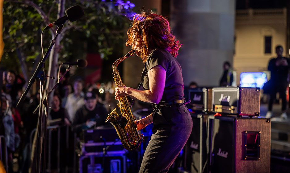 Saxophonist performs at an outdoor concert, surrounded by microphones, amplifiers, and a watching crowd under vibrant purple stage lights.