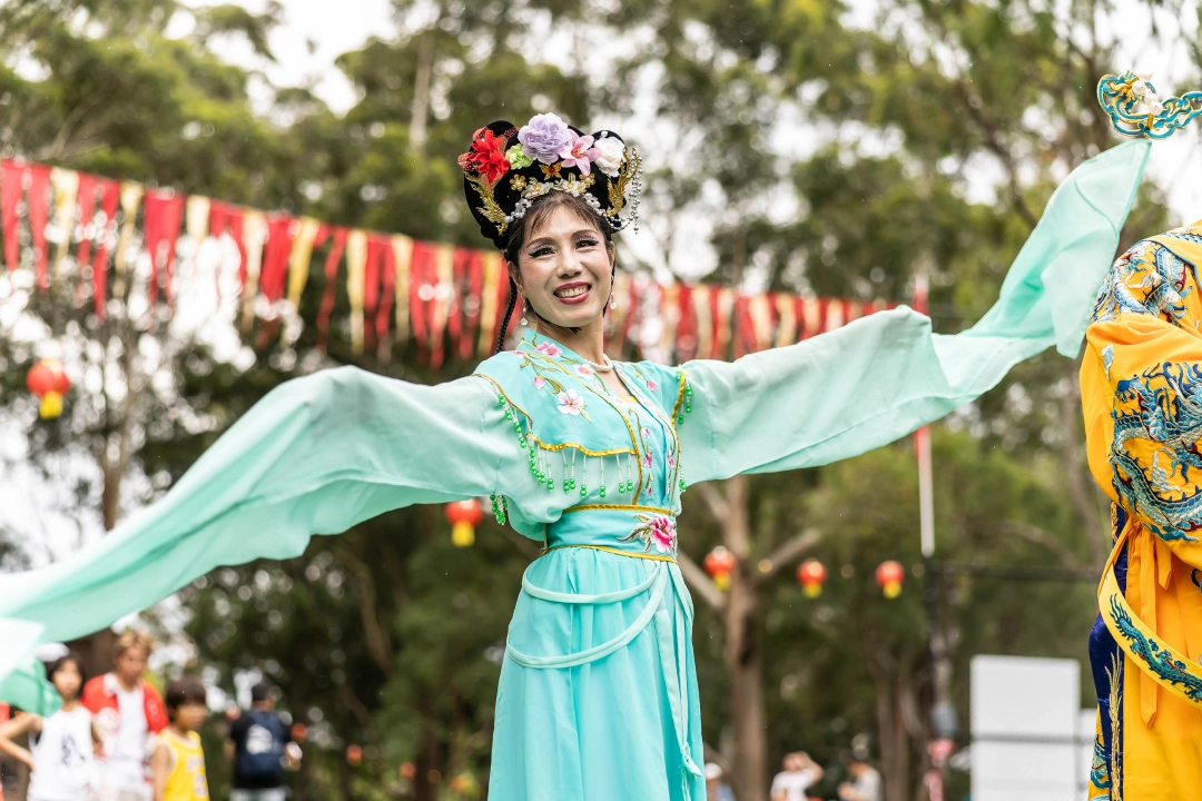 A dancer in a turquoise, flowing costume performs outdoors, surrounded by colorful bunting and trees. The scene is lively and festive.