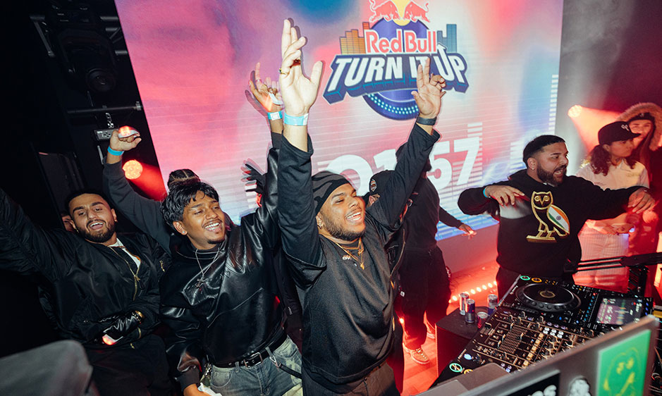 Energetic DJ event with cheering performers raising their hands beneath a Red Bull Turn Up sign, bathed in pink and red stage lights.