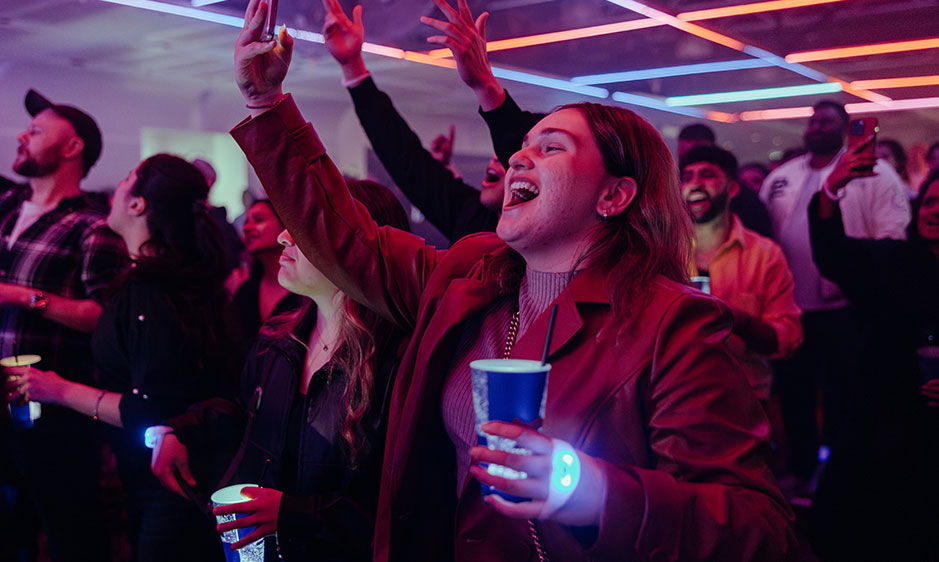 Energetic concertgoers sing and raise their arms beneath colorful neon lights, while a smiling woman in red holds a blue cup.