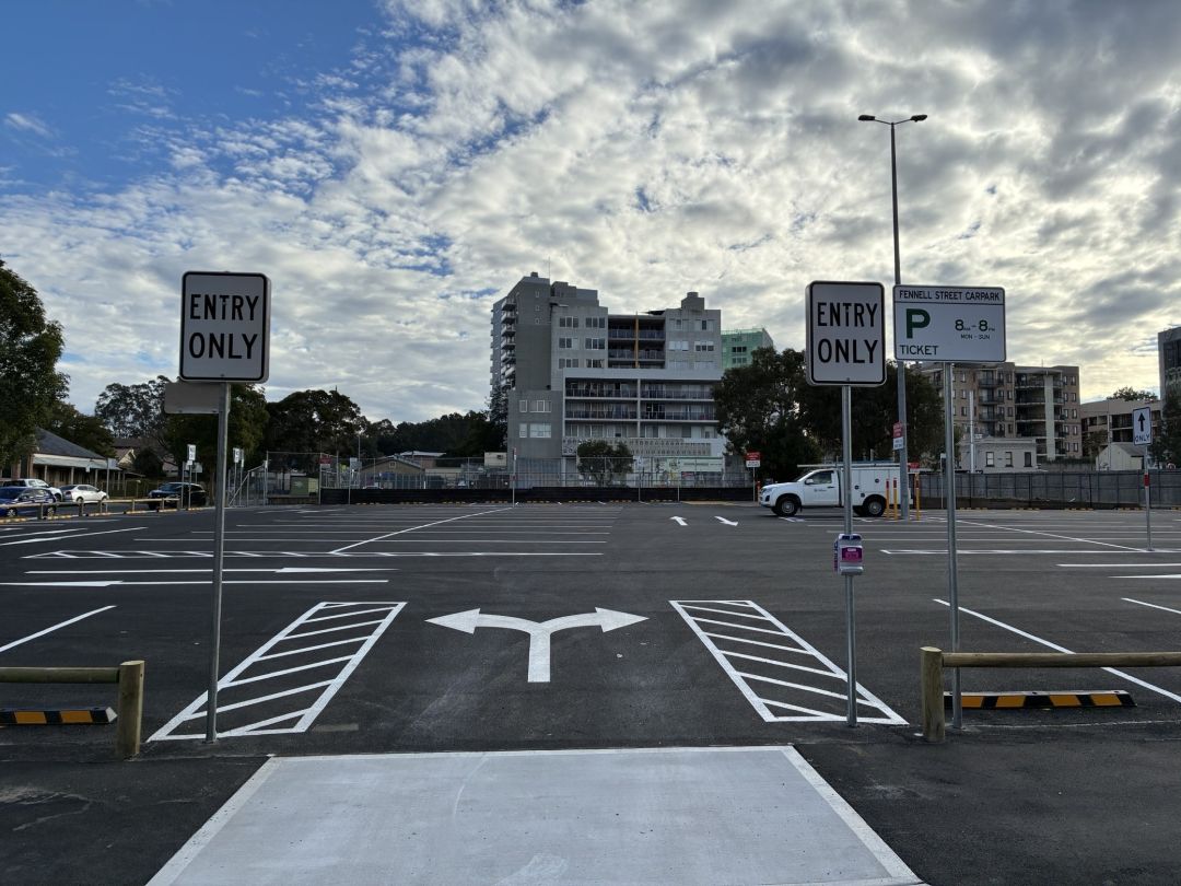 Mostly empty parking lot with ENTRY ONLY signs, directional arrows, marked bays, and high-rise buildings beneath a dramatic cloudy sky.