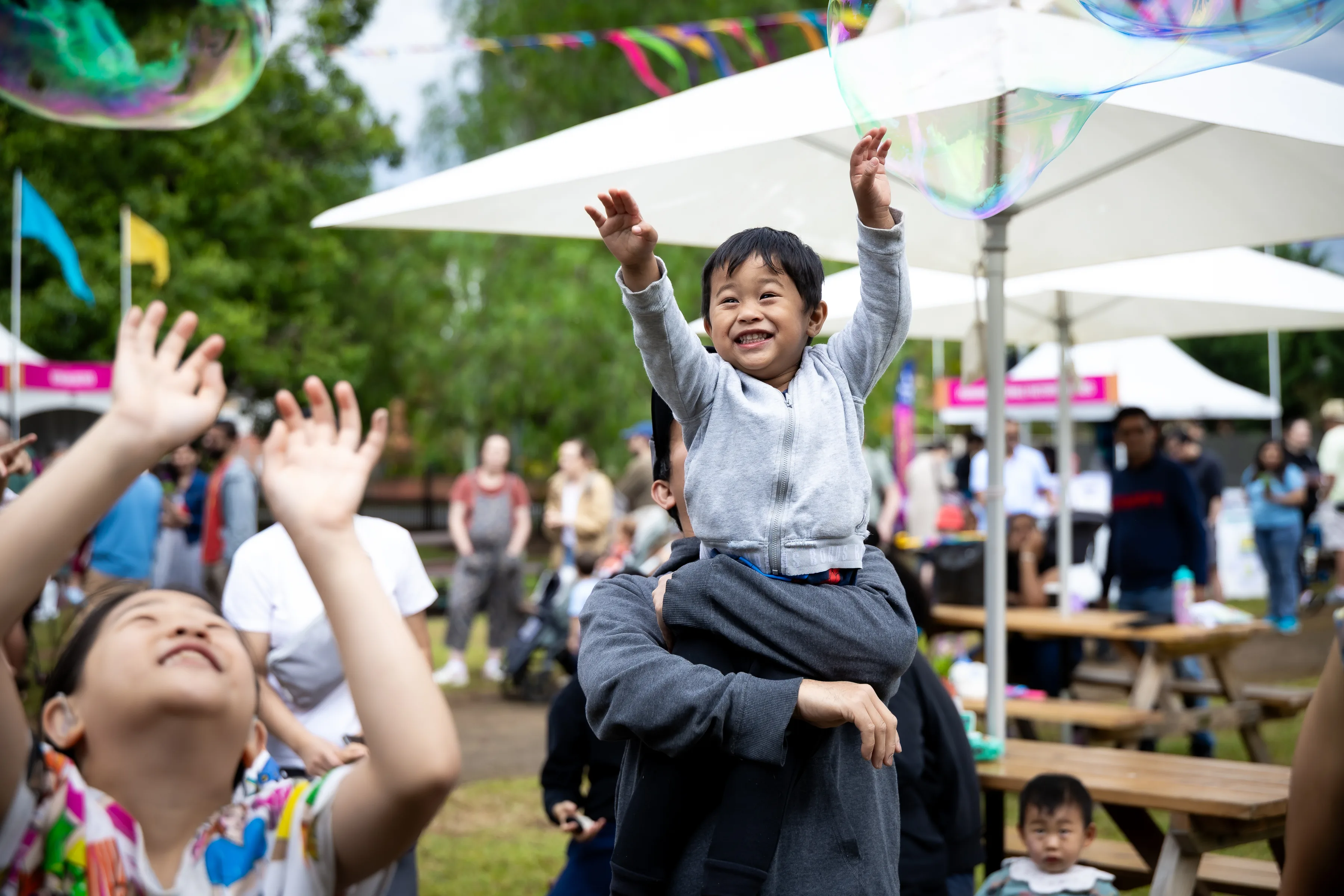 A child sits on an adult's shoulders reaching up toward a large soap bubble at an outdoor community event.