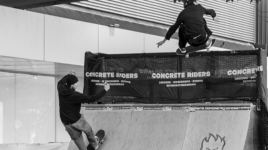 Black-and-white photo of two skateboarders at an indoor half-pipe: one rides up the ramp as another launches above a branded barrier.