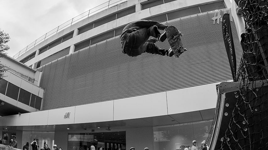 Black-and-white photo of a skateboarder performing an aerial trick beneath a ribbed pedestrian bridge outside a modern shopping center.