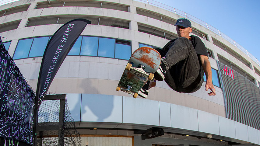 Skateboarder leaps above a ramp outside a shopping mall, gripping a worn skateboard; event banners and an H&M sign frame the action.