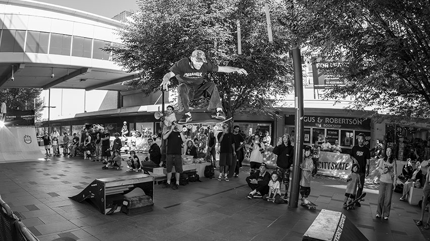 Black-and-white photo of a skateboarder airborne above a ramp in a busy urban plaza, surrounded by spectators and shops; energetic skate event.