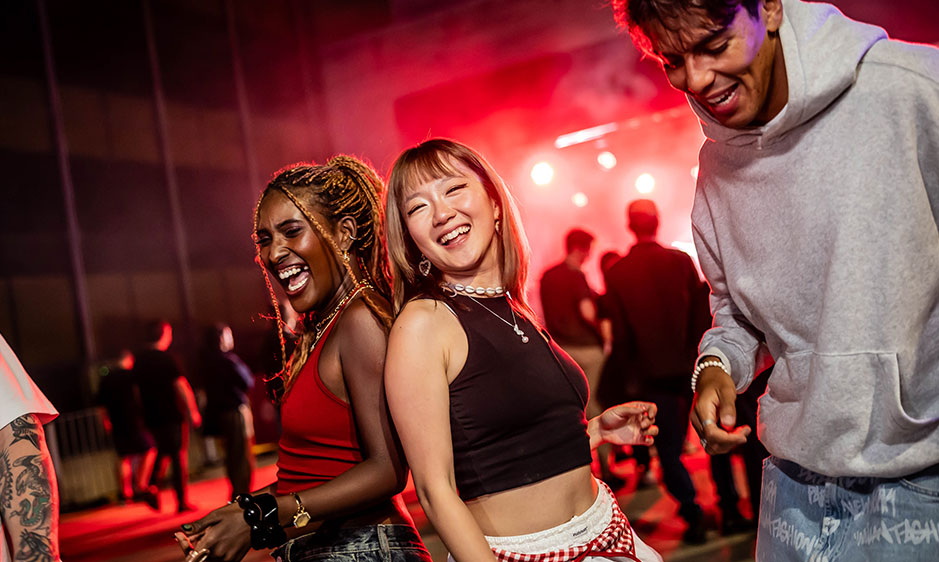 Two women laugh and dance at a lively red-lit party, with blurred guests behind them and a person in a gray hoodie on the right.