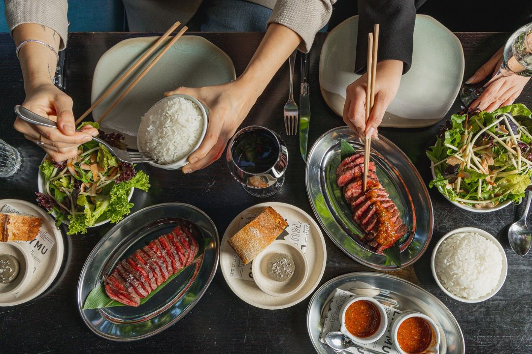 a shared dining table featuring a variety of Asian-inspired dishes, including sliced grilled meat, rice, fresh salads, dipping sauces and desserts.