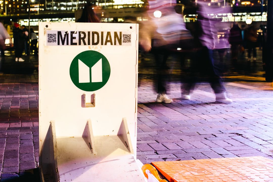 Meridian sign with green logo stands on a city plaza at night, while blurred pedestrians pass behind it, creating a lively, energetic mood.