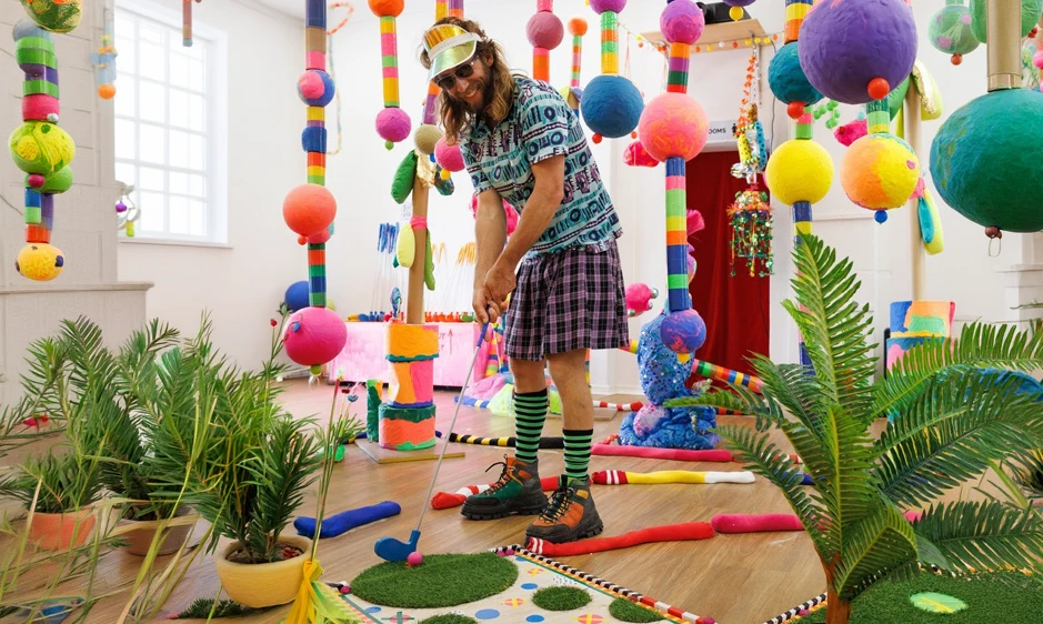 Colorful indoor mini-golf scene with a bearded man putting among tropical plants, hanging pom-poms, and playful decorations; whimsical mood.
