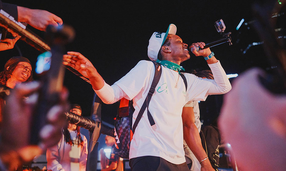 Energetic singer in a white shirt and backward cap performs into a microphone on a dark stage, surrounded by cheering fans and phones.