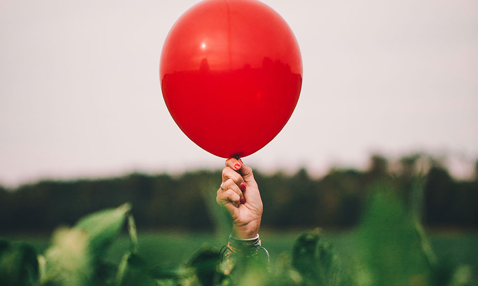 A hand holds a bright red balloon above a green field, with a blurred tree line behind it; the image feels playful, hopeful, and uplifting.