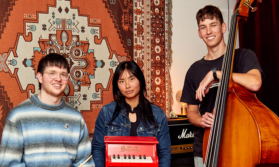 Three smiling musicians pose indoors with a red keyboard: two sit before patterned rugs while a man stands beside an upright bass.