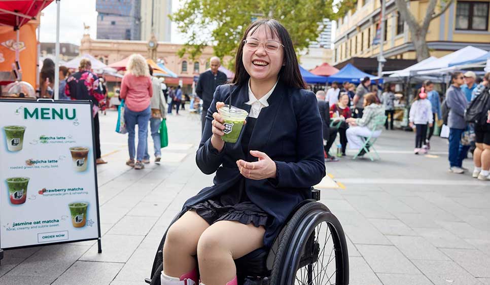 A smiling woman in a wheelchair laughs while holding a green drink at a busy outdoor market, with colorful stalls and a menu board behind her.