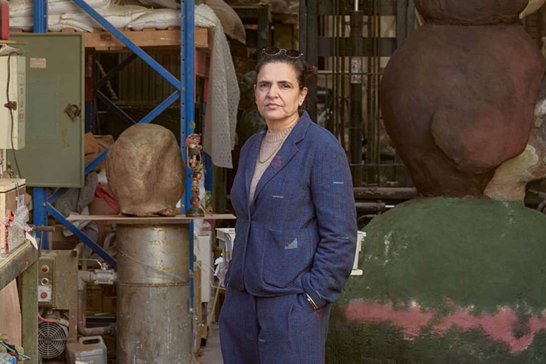 A composed woman in a blue suit stands in a cluttered sculpture workshop, surrounded by oversized painted heads, tools, shelves, and metal frames.