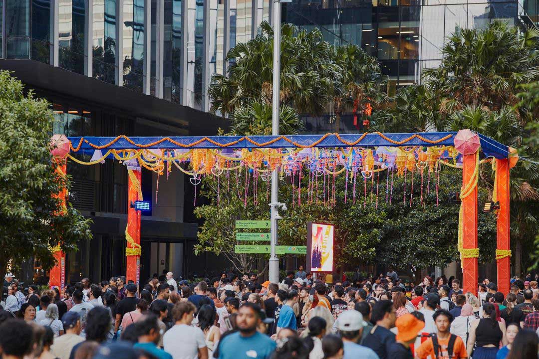 A large crowd gathers beneath a colorful festival arch outside modern glass buildings, creating a lively, festive street scene in the city.
