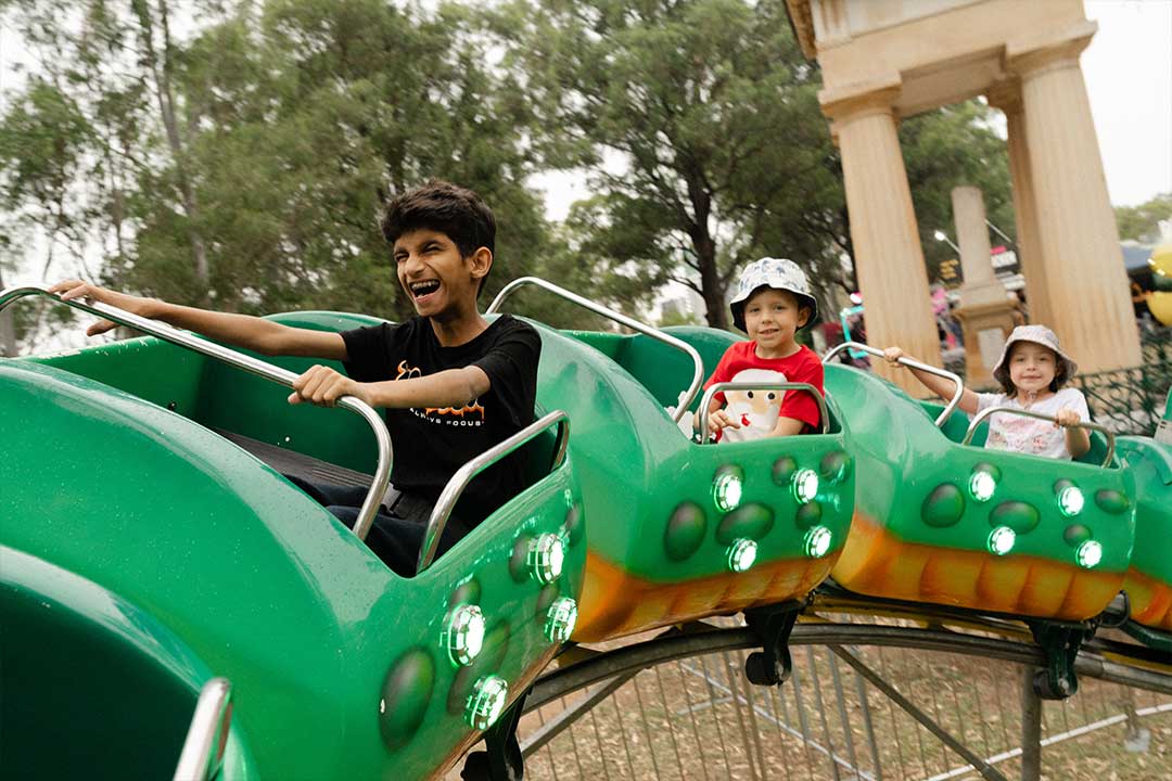 A man and two children laugh aboard bright green, light-up amusement park cars, with leafy trees and a columned structure behind them.