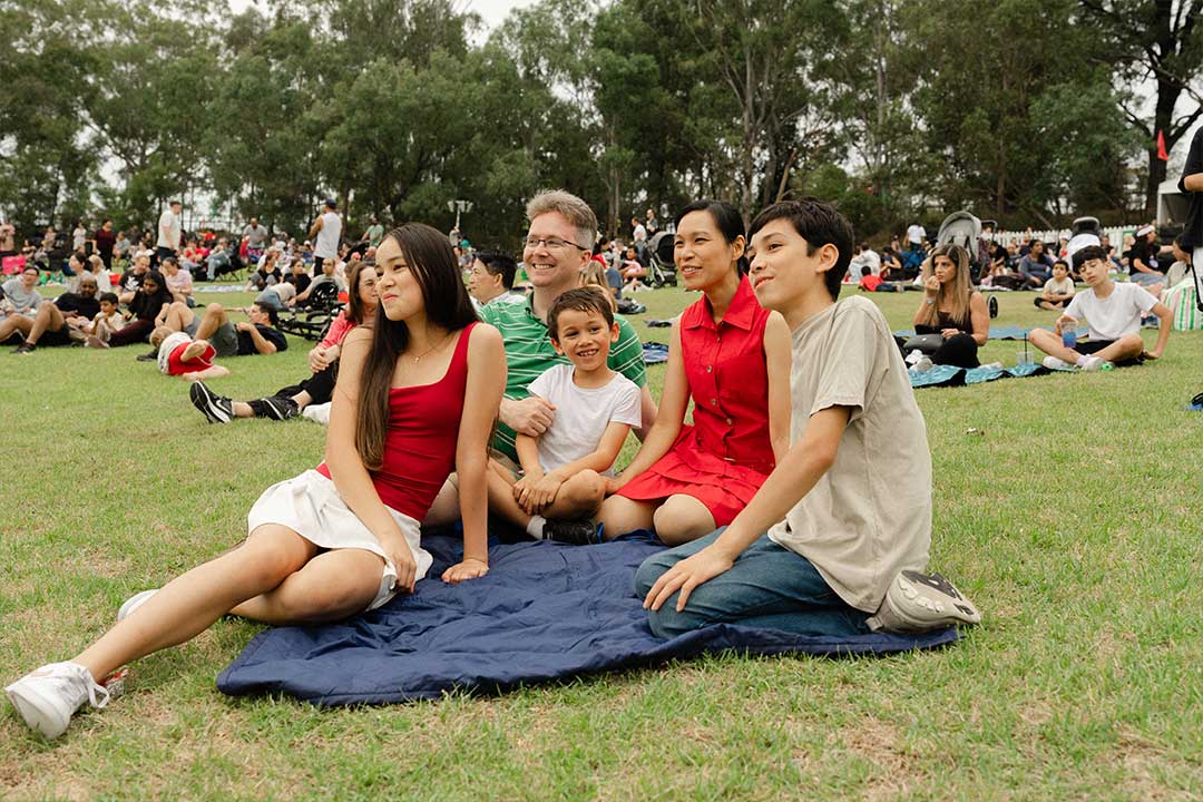 A family of five smiles for a photo on a picnic blanket at a crowded outdoor park gathering, surrounded by trees and relaxed festivalgoers.