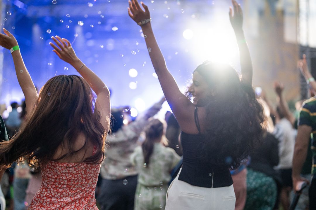 Women dance with raised arms at an outdoor concert as bubbles drift through blue stage lights, creating a joyful, carefree festival atmosphere.
